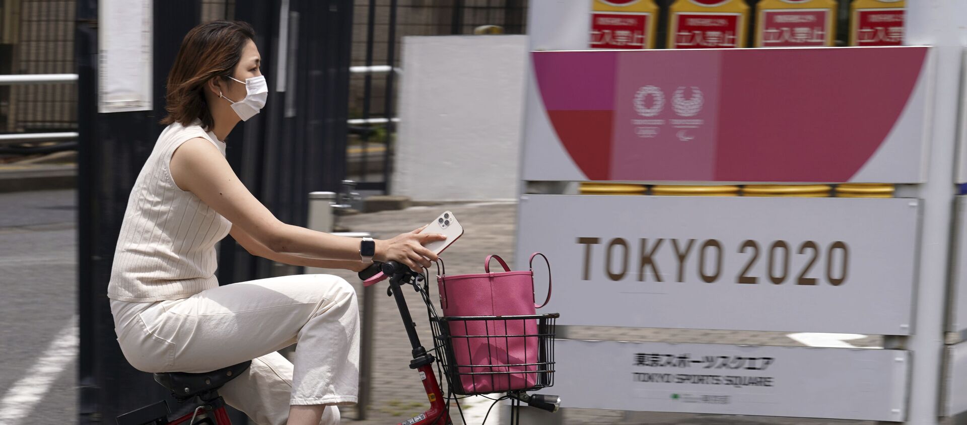 Женщина в защитной маске проезжает на велосипеде мимо баннера Олимпийских и Паралимпийских игр в Токио  - Sputnik 日本, 1920, 22.07.2021